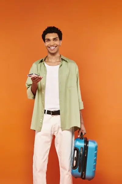 Joven alegre afroamericano hombre en traje acogedor sosteniendo teléfono y maleta y mirando a la cámara - foto de stock