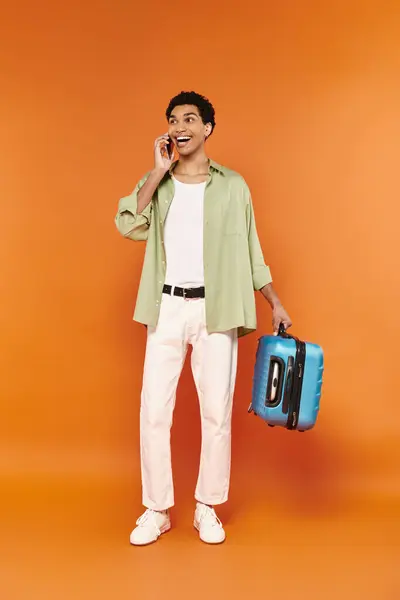 Atractivo jolly africano americano hombre en acogedor traje sosteniendo maleta y hablando por teléfono móvil - foto de stock