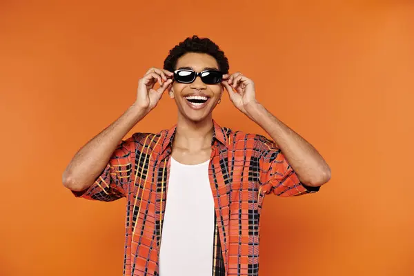 Fröhliche junge afrikanisch-amerikanische Mann in lebendiger Kleidung mit Sonnenbrille posiert auf orangefarbenem Hintergrund — Stockfoto