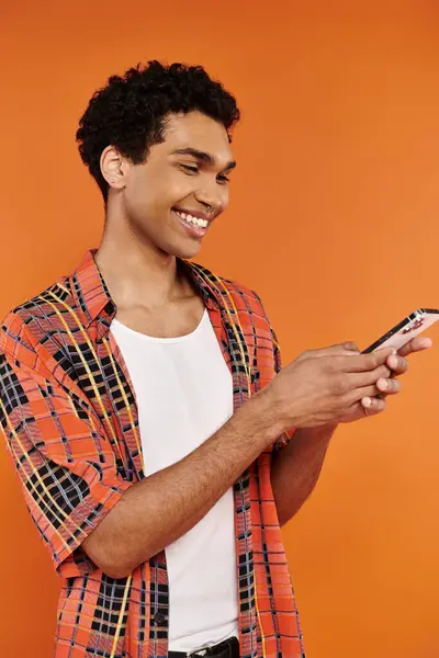 Elegante gioioso uomo africano americano in abiti vivaci guardando il suo telefono su sfondo arancione — Foto stock