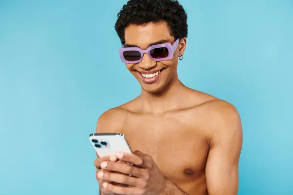 Joyous african american man with stylish sunglasses in swimming trunks holding his smartphone — Stock Photo