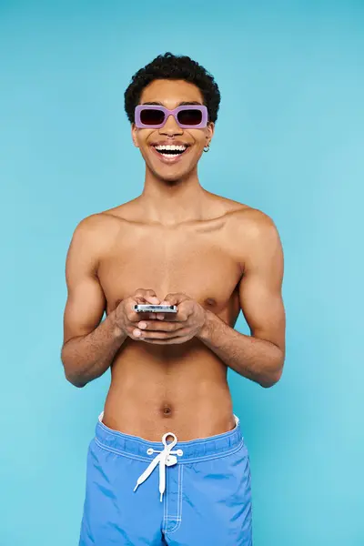 Joyful african american man with stylish sunglasses in swimming trunks holding his smartphone — Stock Photo
