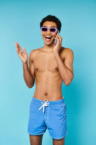 Alegre hombre afroamericano bien parecido en bañador troncos con gafas de sol con estilo hablando por teléfono - foto de stock
