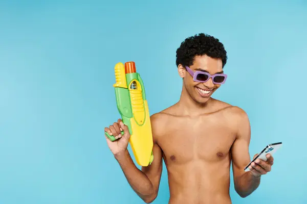 Hombre americano africano alegre con gafas de sol elegantes que sostienen el teléfono y la pistola de agua en telón de fondo azul - foto de stock