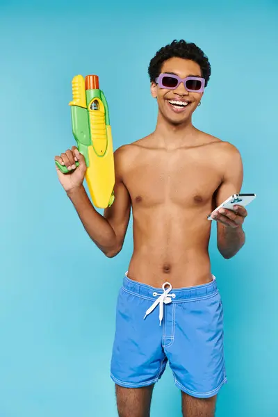 Joyous african american man with stylish sunglasses holding phone and water gun on blue backdrop — Stock Photo