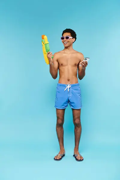 Hombre americano africano alegre con gafas de sol con estilo sosteniendo teléfono y pistola de agua en telón de fondo azul - foto de stock