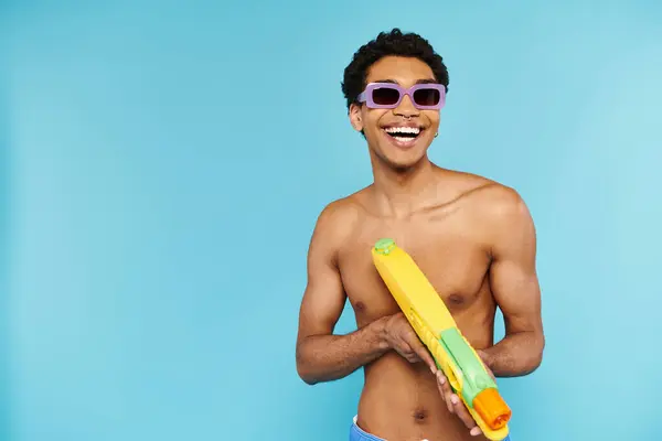 Positivo joven afroamericano hombre en traje de baño con gafas de sol elegantes posando con pistola de agua - foto de stock