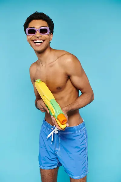 Upbeat joven afroamericano hombre en traje de baño con gafas de sol elegantes posando con pistola de agua - foto de stock