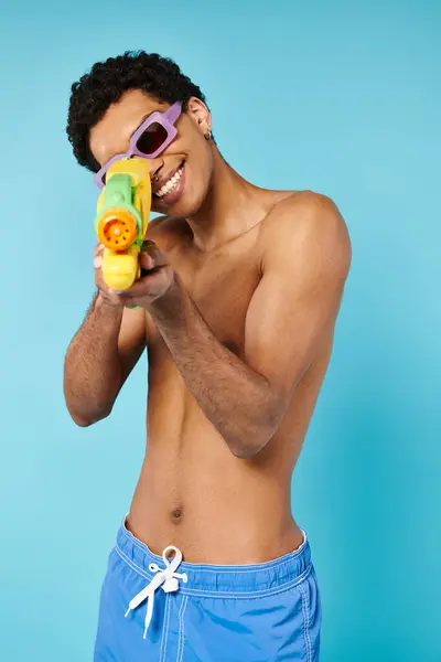 Hombre afroamericano satisfecho en bañador con gafas de sol elegantes posando con pistola de agua - foto de stock