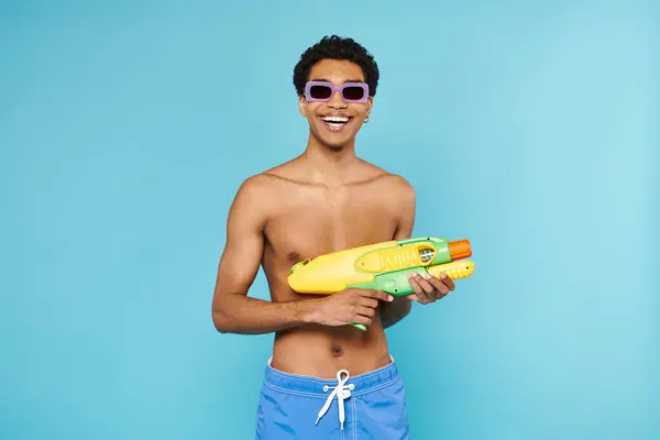 Joyful young african american man in swimming trunks with stylish sunglasses posing with water gun — Stock Photo
