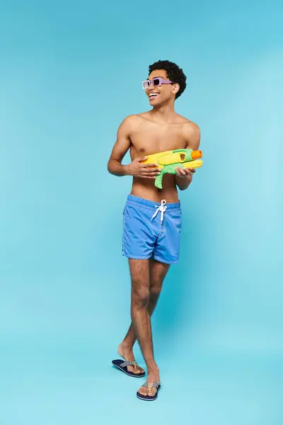 Joyous young african american man in swimming trunks with stylish sunglasses posing with water gun — Stock Photo