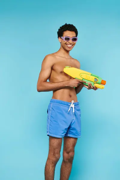 Alegre joven afroamericano hombre en traje de baño con gafas de sol elegantes posando con pistola de agua - foto de stock