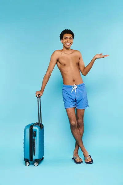 Alegre joven afroamericano en traje de baño posando junto a la maleta y sonriendo a la cámara - foto de stock