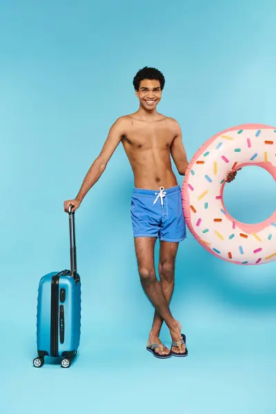 Alegre afroamericano hombre en traje de baño con anillo de natación y maleta sonriendo a la cámara - foto de stock