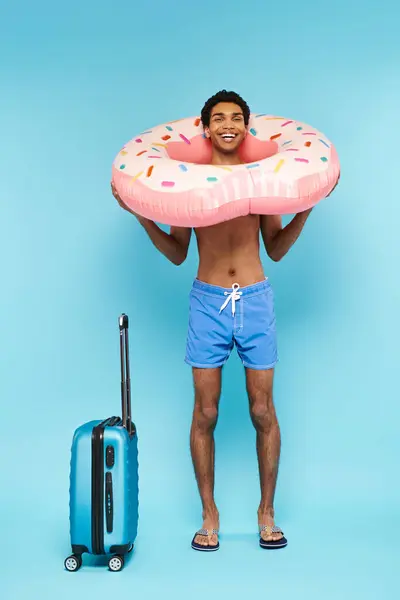Merry african american man in swimming trunks with swimming ring and suitcase smiling at camera — Stock Photo