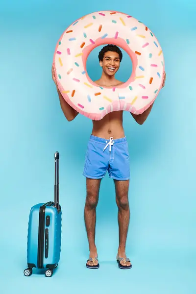 Hombre afroamericano positivo en pantalones de baño con anillo de natación y maleta sonriendo a la cámara - foto de stock