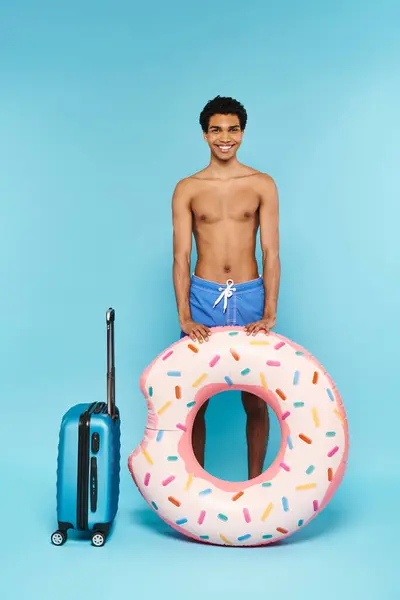 Alegre afroamericano hombre en traje de baño con anillo de natación y maleta sonriendo a la cámara - foto de stock