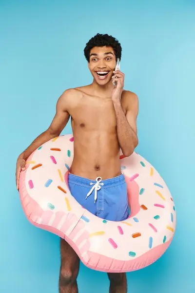 Gut aussehende fröhliche afrikanisch-amerikanische Mann in Badehose mit aufblasbarem Donut telefoniert — Stockfoto