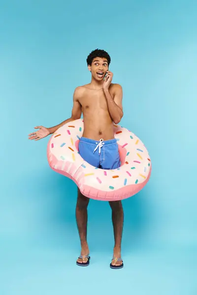 Beau homme afro-américain joyeux dans les troncs de natation avec beignet gonflable parlant par téléphone — Photo de stock