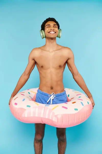 Hombre americano africano alegre en los baúles de natación con la rosquilla inflable que disfruta de la música en auriculares - foto de stock