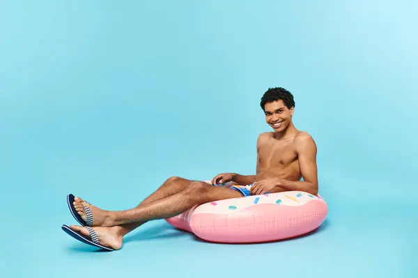 Alegre afroamericano hombre en bañador troncos sentado en inflable donut y sonriendo a cámara - foto de stock