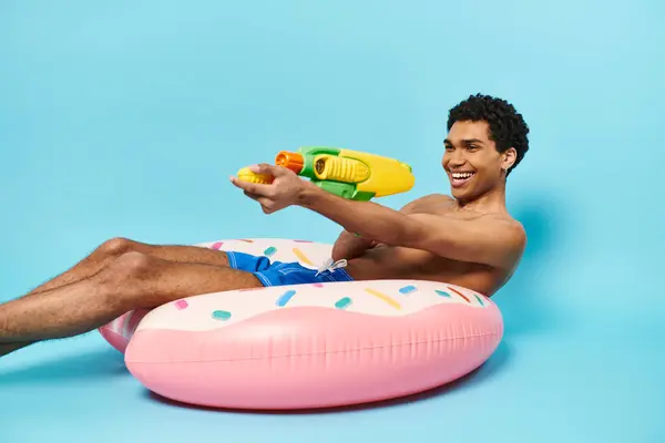 Optimista atractivo hombre afroamericano posando con pistola de agua en donut inflable sobre fondo azul - foto de stock