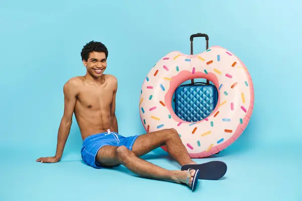 Positivo joven afroamericano hombre en bañador troncos sentado cerca de la maleta y donut inflable - foto de stock