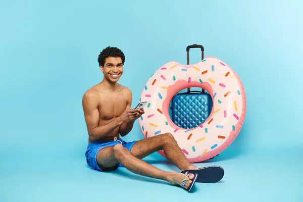 Alegre afroamericano hombre sosteniendo su teléfono inteligente y sentado cerca de la maleta y donut inflable - foto de stock