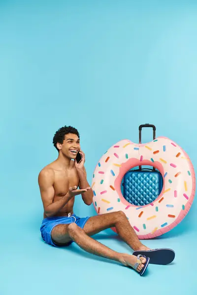 Hombre americano africano alegre que habla por el teléfono móvil al lado de su maleta y donut inflable - foto de stock