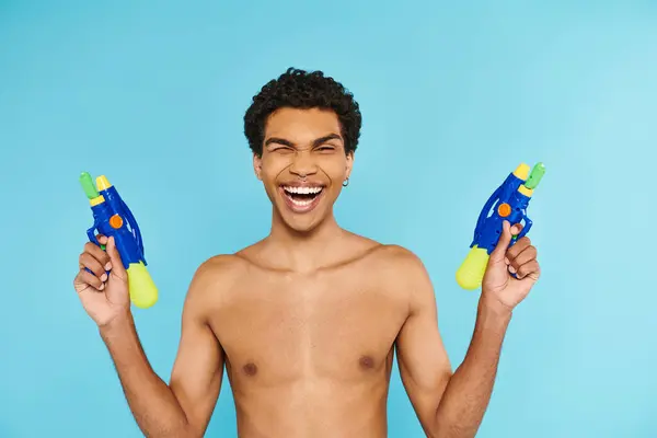 Positivo guapo afroamericano hombre posando con dos pistolas de agua y sonriendo a la cámara felizmente - foto de stock