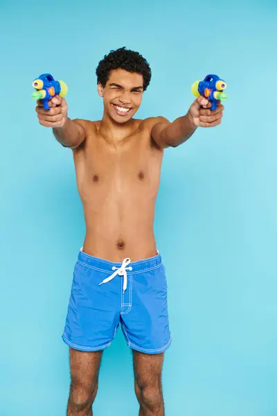 Alegre guapo afroamericano hombre posando con dos pistolas de agua y sonriendo a la cámara felizmente - foto de stock
