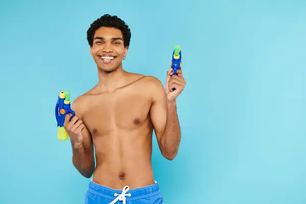 Alegre guapo afroamericano hombre posando con dos armas de agua y sonriendo a la cámara felizmente - foto de stock