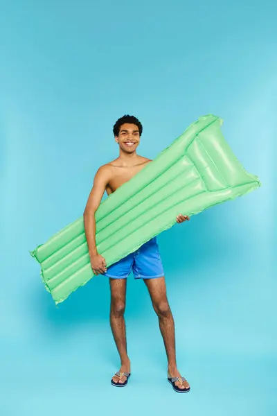Alegre afroamericano hombre en traje de baño posando con colchón de aire y sonriendo a la cámara - foto de stock