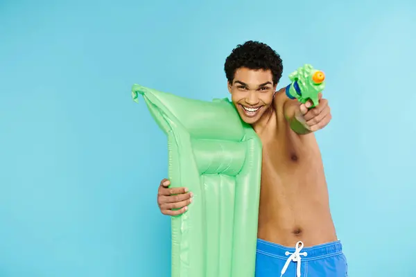 Cheerful young african american man posing with air mattress and water gun on blue background — Stock Photo
