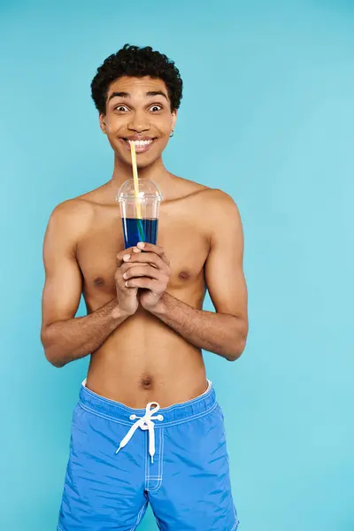 Alegre afroamericano hombre en bañador troncos disfrutando refrescante cóctel y sonriendo a la cámara - foto de stock