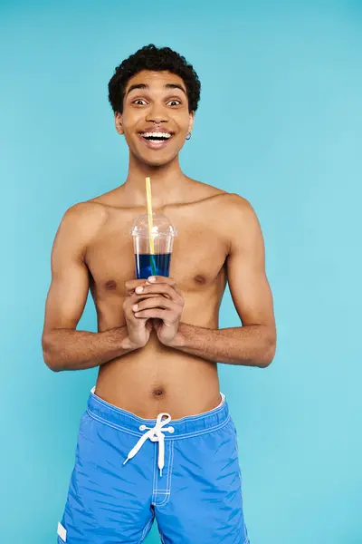 Alegre afroamericano hombre en bañador troncos disfrutando refrescante cóctel y sonriendo a la cámara — Stock Photo