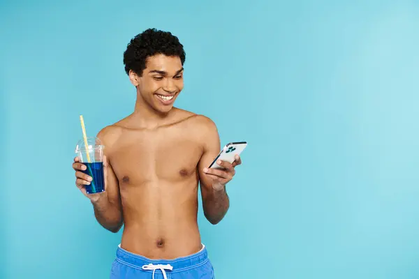 Alegre afroamericano hombre en bañador troncos mirando su teléfono y la celebración de cóctel en la mano - foto de stock
