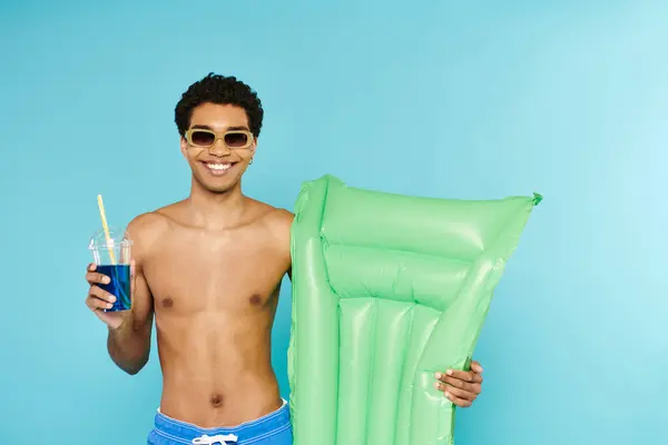 Alegre afroamericano hombre disfrutando refrescante cóctel celebración colchón de aire sobre fondo azul - foto de stock