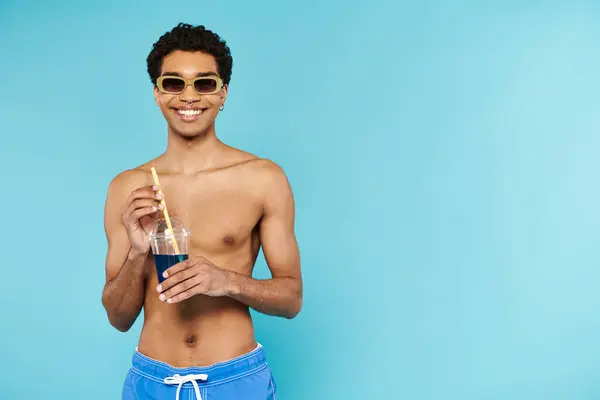 Handsome jolly african american man in swimming trunks with sunglasses enjoying refreshing cocktail — Stock Photo