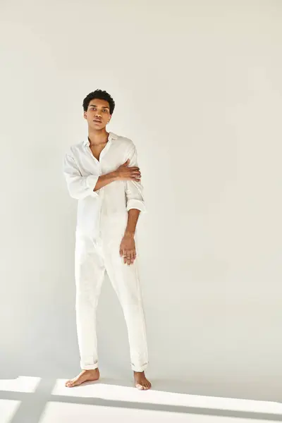 Elegante hombre afroamericano en traje blanco posando descalzo y mirando a la cámara sobre fondo beige - foto de stock