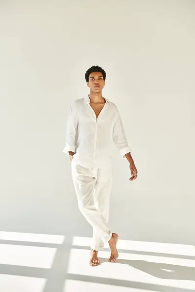 Young african american man in white attire posing barefoot and looking at camera on beige backdrop — Stock Photo