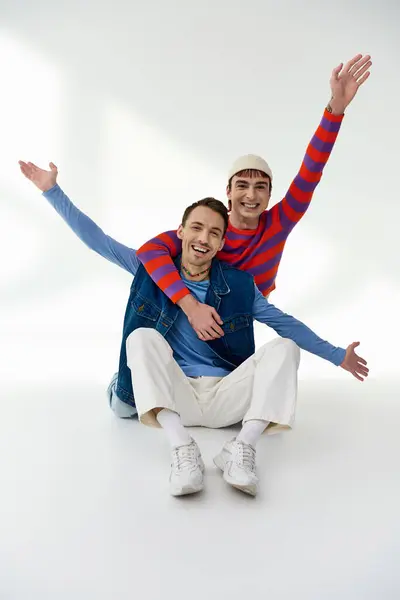 Dos amigos lgbt con estilo feliz en trajes vibrantes mirando a la cámara y posando sobre fondo gris - foto de stock