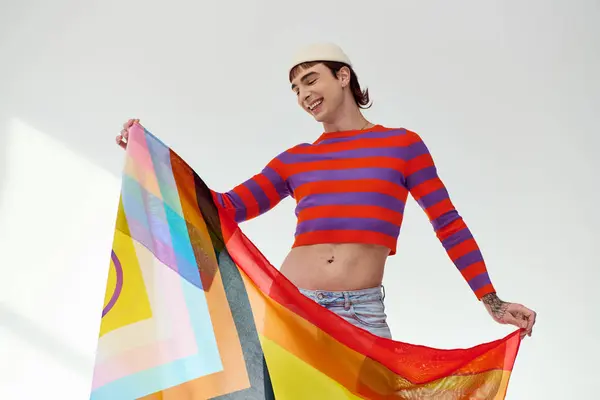 Молодий привабливий гей-чоловік у яскравому вбранні позує з веселковим прапором і дивиться на сірий фон — стокове фото