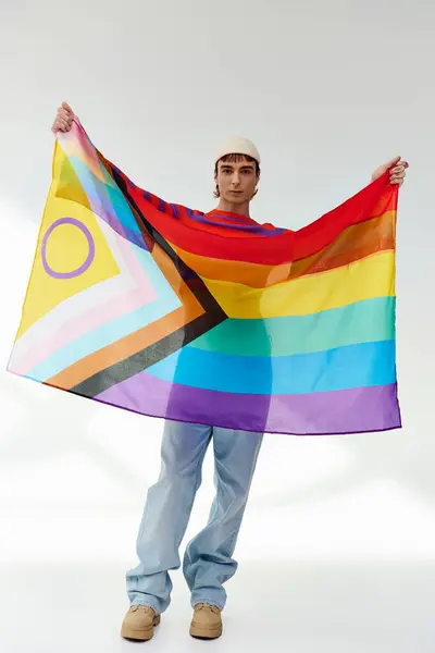 Радісний привабливий гей чоловік у яскравому вбранні з білим капелюхом, що тримає веселковий прапор і дивиться на камеру — стокове фото