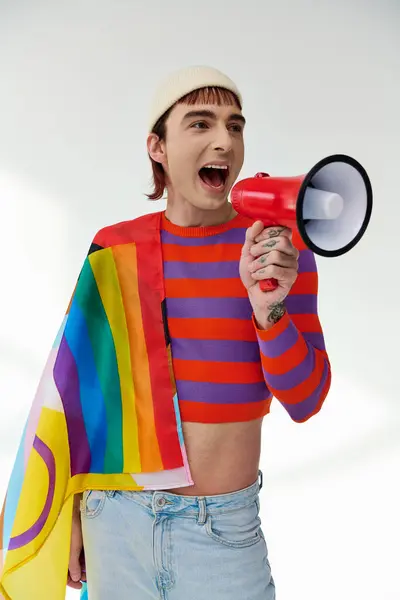 Jolly stylish gay man in vibrant attire with rainbow flag using megaphone and looking away — Stock Photo