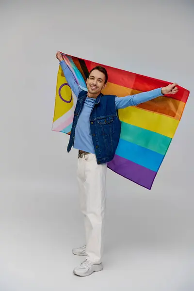 Positivo atractivo gay hombre en vibrante casual atuendo celebración arco iris bandera y sonriendo a la cámara - foto de stock