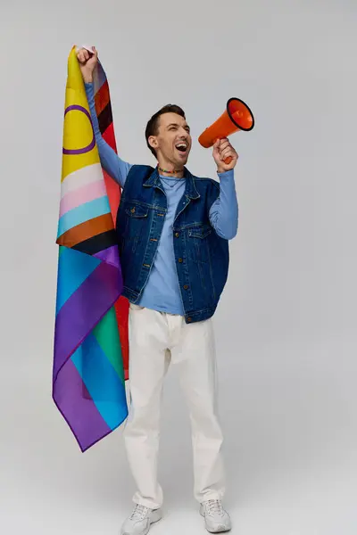 Joven bien parecido gay hombre en acogedor atuendo sosteniendo arco iris bandera y el uso de megáfono en gris telón de fondo - foto de stock