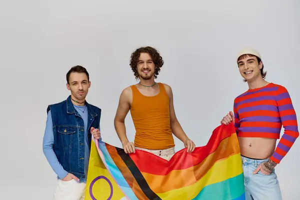 Tres alegre guapo gay los hombres en vibrante ropa posando con arco iris bandera y mirando cámara - foto de stock