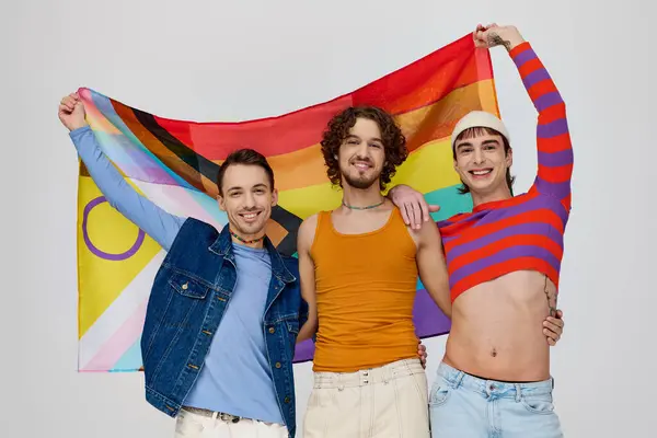 Three joyful handsome gay men in vibrant clothes posing with rainbow flag and looking at camera — Stock Photo