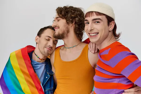 Три радісні стильні геї в затишному одязі позують активно з веселковим прапором на сірому фоні — стокове фото